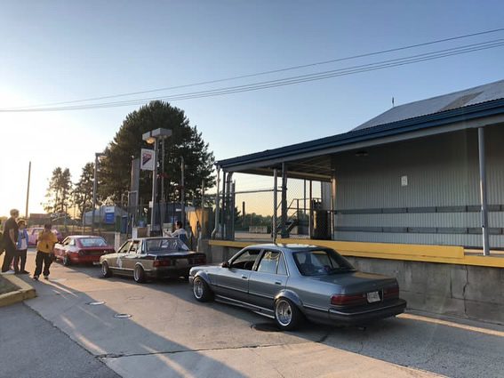 Vintage cars lined up near a gas station on a sunny day with a group of people in the background.