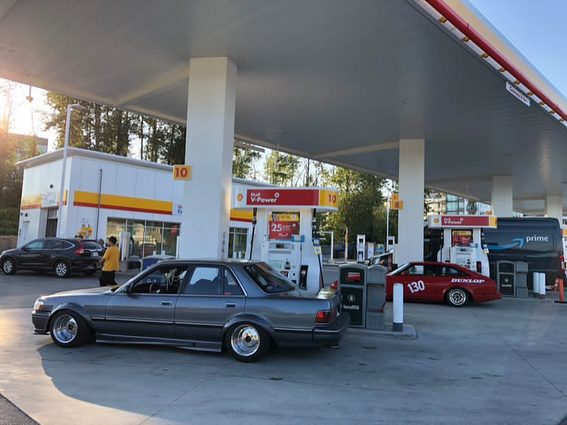 Classic and modern cars refueling at a Shell gas station under a clear sky.