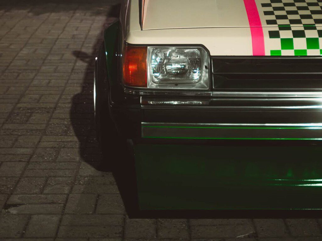Close-up of vintage car front with pink and green checkered design on hood, parked on brick pavement at night.