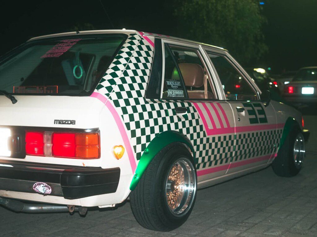 Vintage Mazda with custom checkered and pink design parked at night.