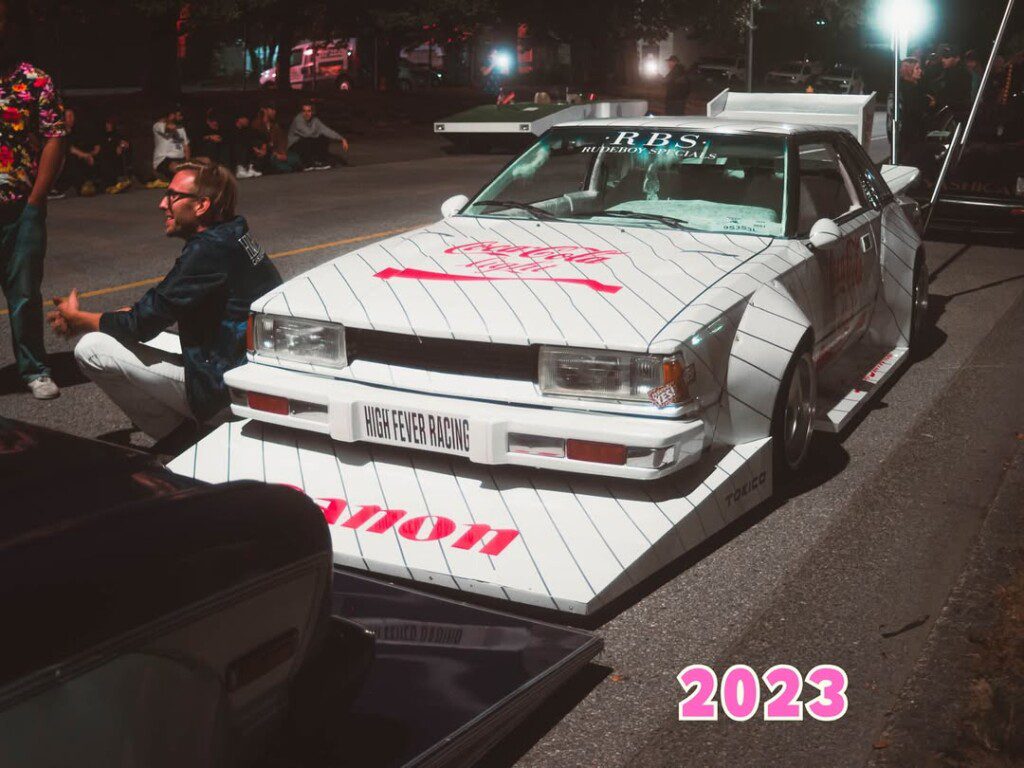 Custom retro car with striking decals at a nighttime car show in 2023.