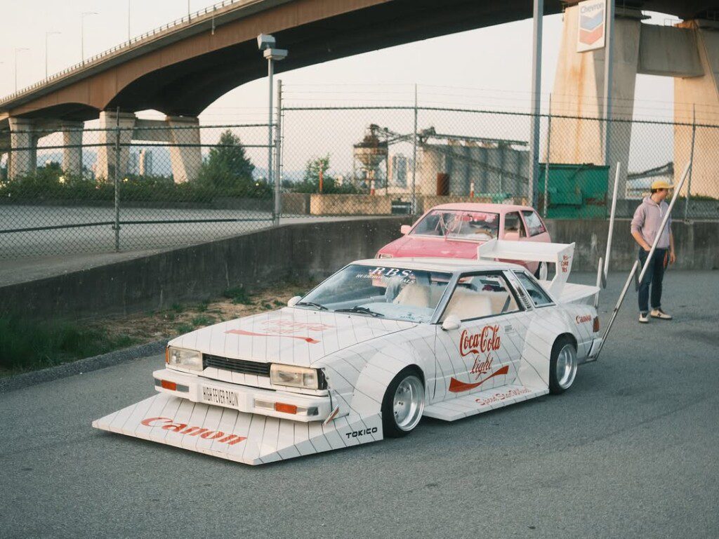 Custom retro cars with bold graphics parked under a bridge, featuring unique body kits and vintage styles.