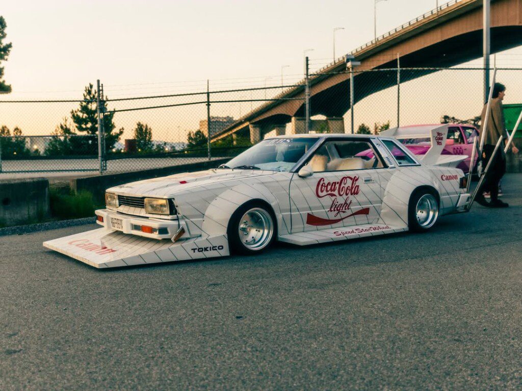 Customized retro car with Coca-Cola branding parked by a fence at sunset, showcasing unique body modifications.