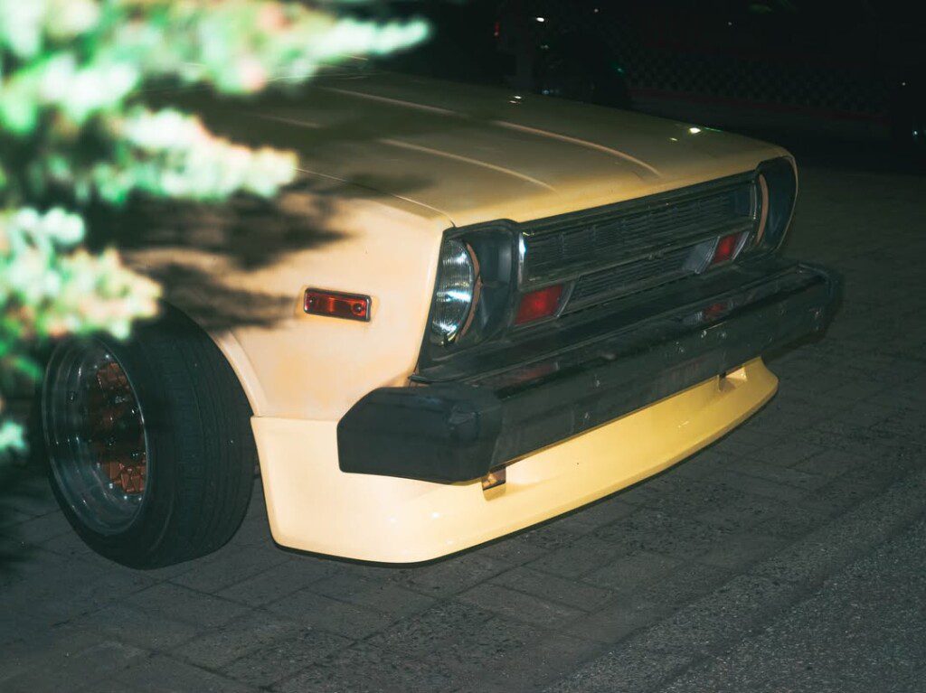 Yellow vintage car parked on a dimly lit street at night, partially obscured by tree branches.