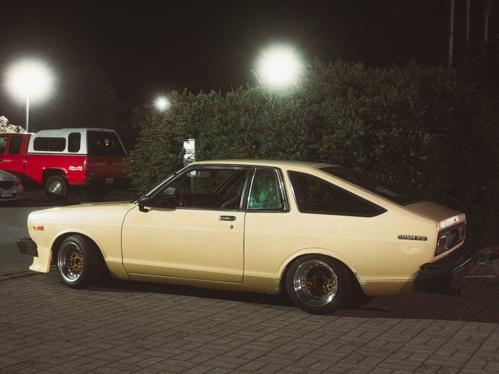 Vintage yellow Datsun 210 coupe parked at night, illuminated by streetlights.