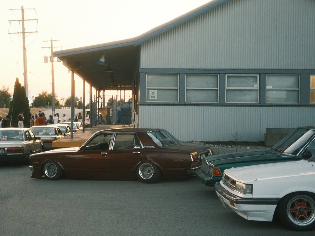 Vintage cars parked at an industrial building during sunset, with a group of people in the background.