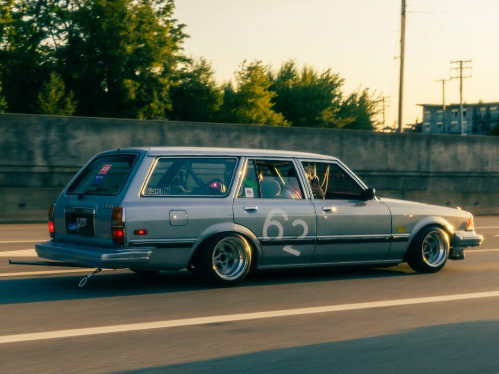Classic silver station wagon with custom wheels and decals driving on highway at sunset.