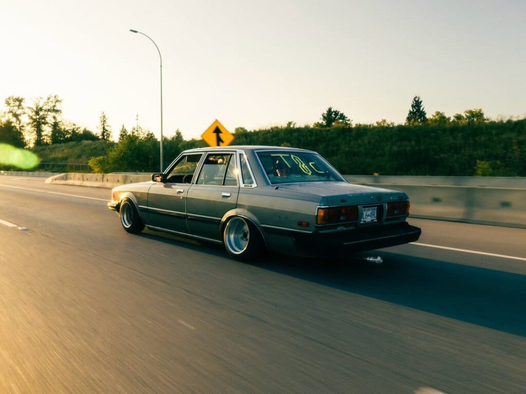 Vintage car cruising on a sunlit highway, surrounded by greenery and a clear sky.