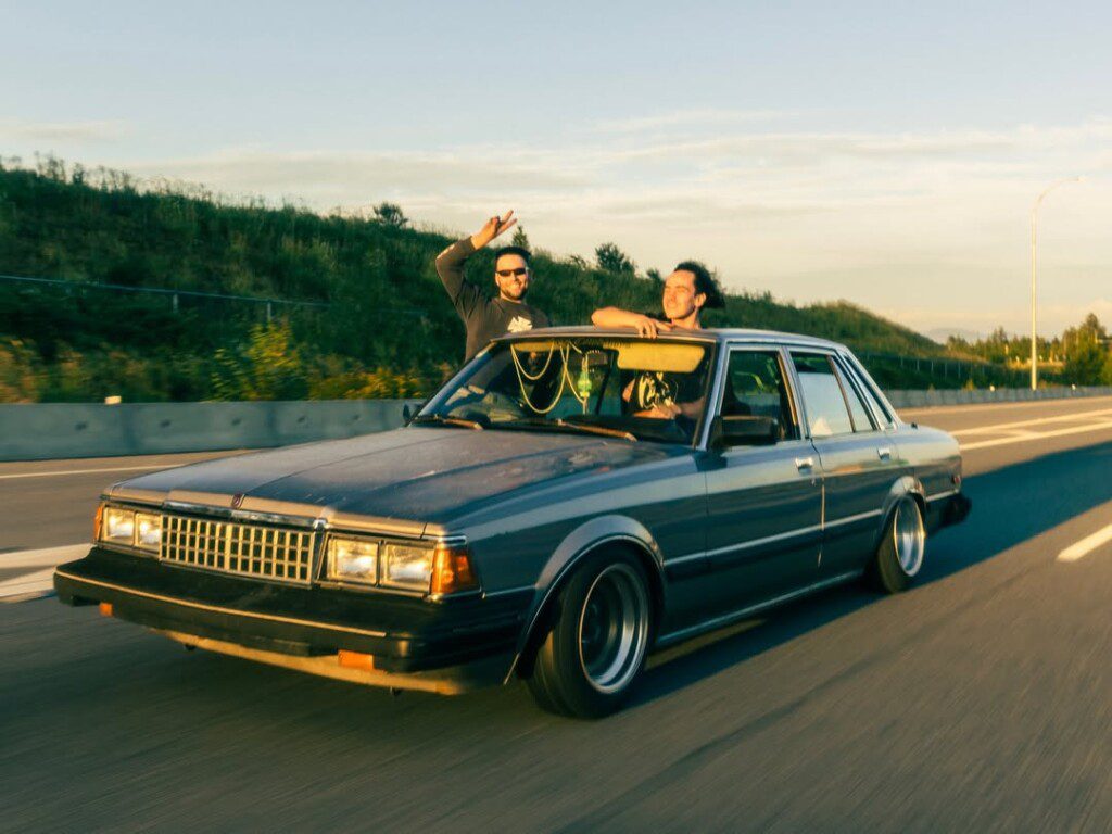 Vintage car cruising on highway with two passengers waving and enjoying a sunny day.
