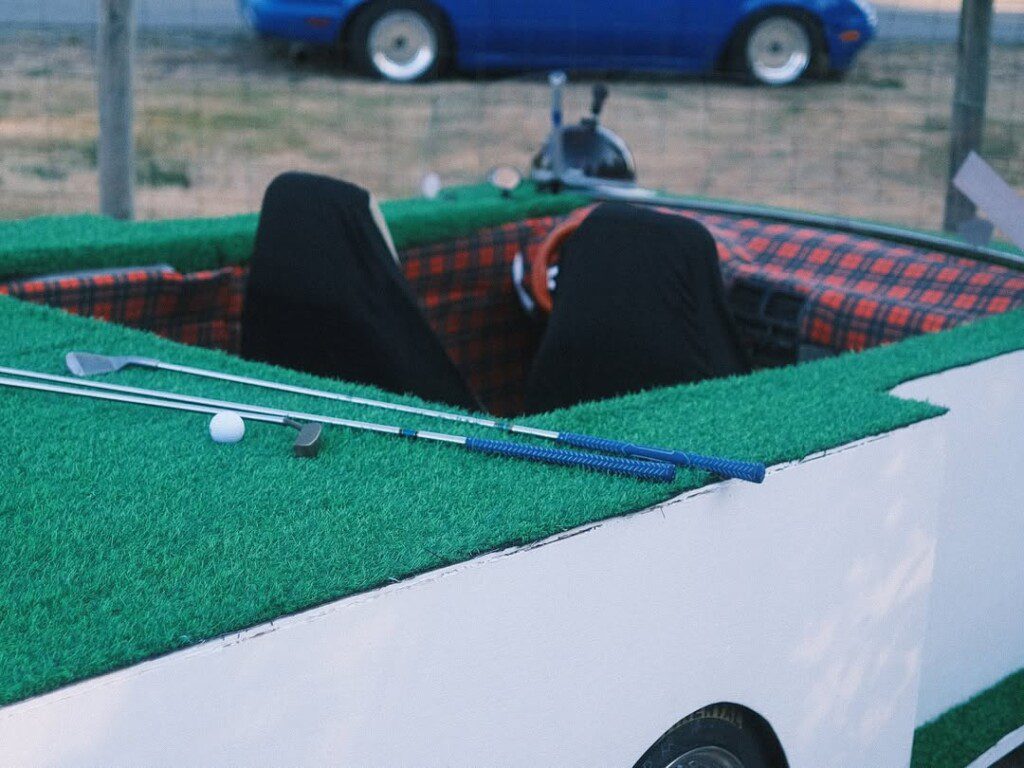 Golf-themed car with turf, two golf clubs, and a ball on the roof. Unique vehicle with checkered interior.
