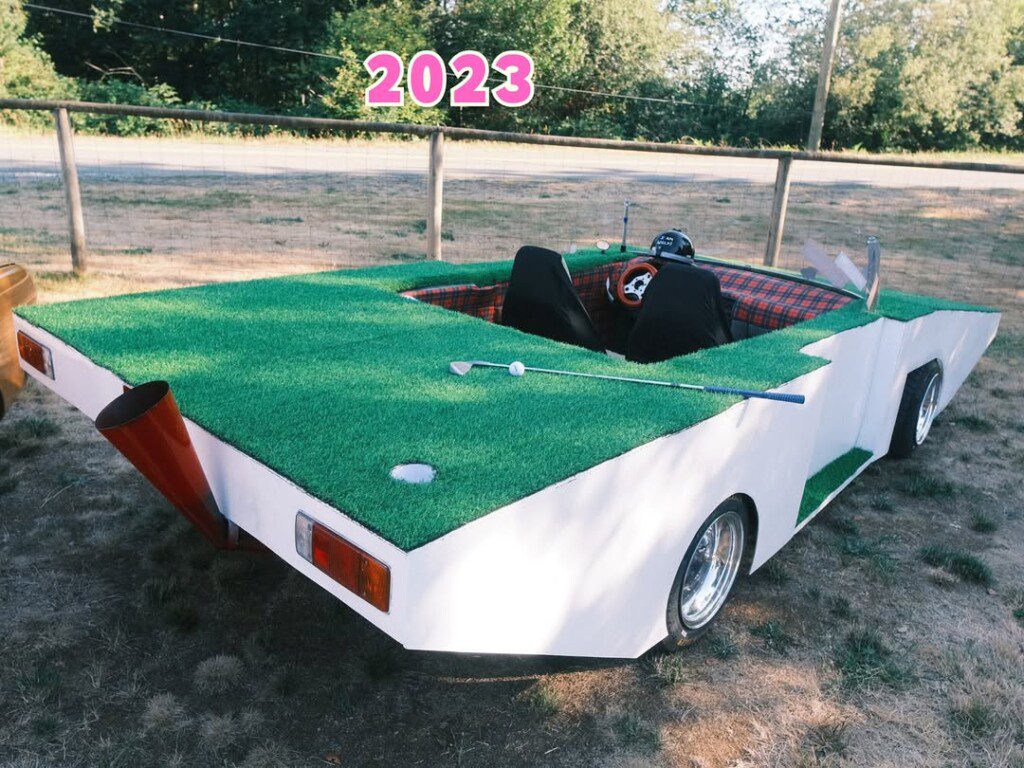 Futuristic golf-cart car with grass design and 2023 text, parked outdoors in a rustic setting.