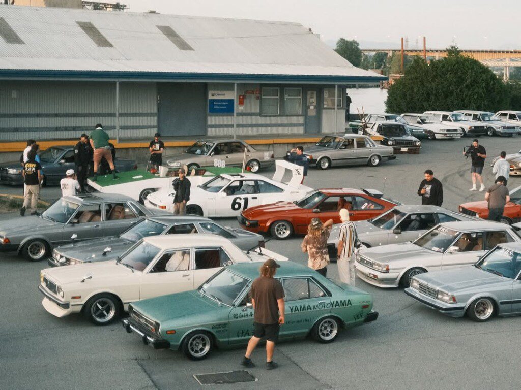 Vintage car meet with various classic models and enthusiasts gathered in a parking lot near an industrial building.