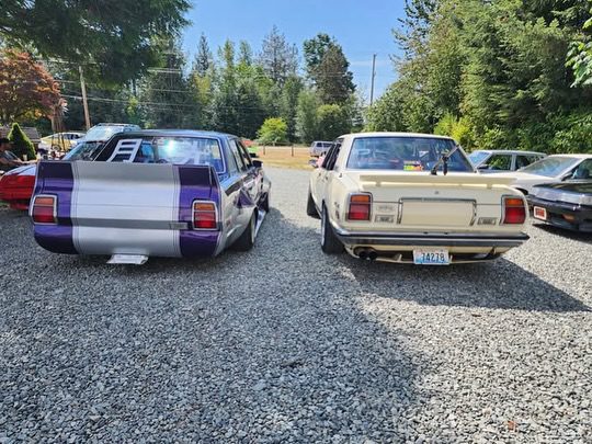 Two vintage cars with custom paint parked on gravel, surrounded by trees on a sunny day.