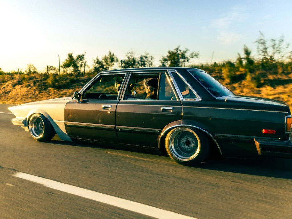 Classic vintage car driving on a sunny road, side view with trees in the background.
