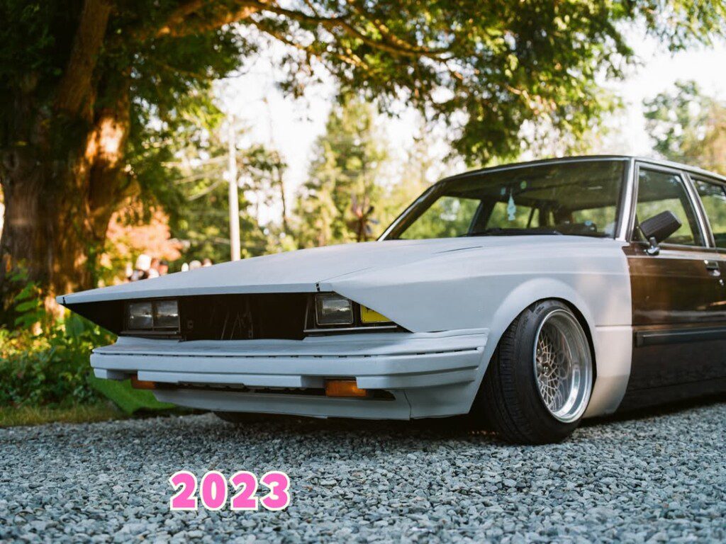 Sleek vintage car parked on gravel with trees in the background, featuring bold 2023 text.