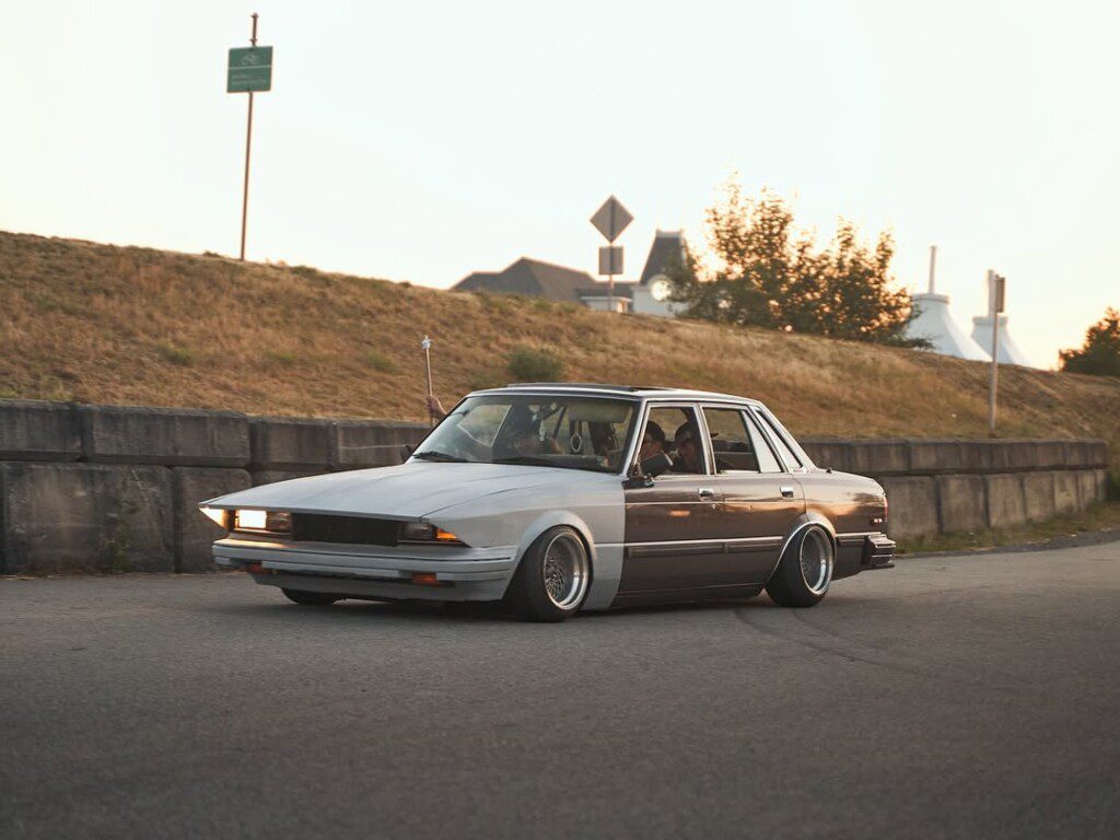 Classic modified sedan cruising on a scenic road at sunset, showcasing low stance and custom wheels.