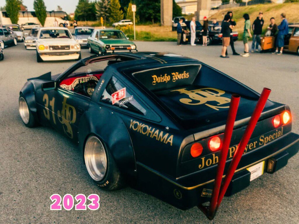 Custom retro car with bold decals at a car meet, featuring metal exhaust pipes, surrounded by classic cars, 2023.