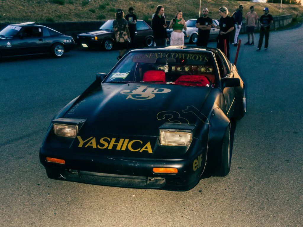Modified black sports car with Yashica logo at a car meet, surrounded by people and other vehicles.