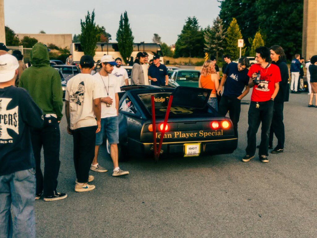 Group gathers around a vintage sports car labeled Icon Player Special at a car meet in an open parking lot.