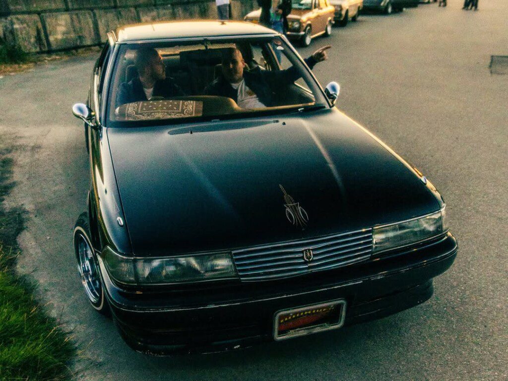 Black classic car with two passengers on street, driver pointing, other vintage cars in background, evening setting.