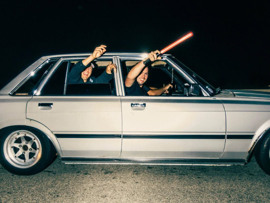 Two people in a vintage car at night, smiling and waving a red light stick.