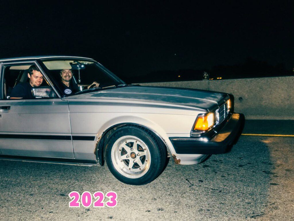 Two people smiling in a vintage car at night with 2023 in pink lettering on the road.