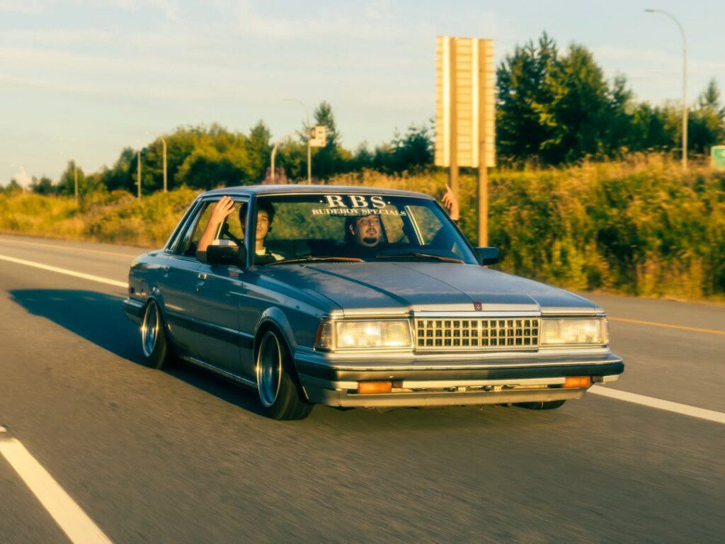 Classic blue car cruising down highway in sunlight, vintage style, with scenic greenery in background.