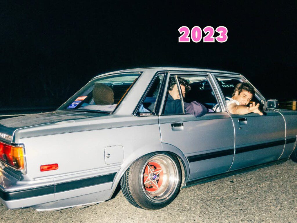 Vintage car cruise at night, passengers enjoying the ride, vibrant 2023 text above. Retro vibes.