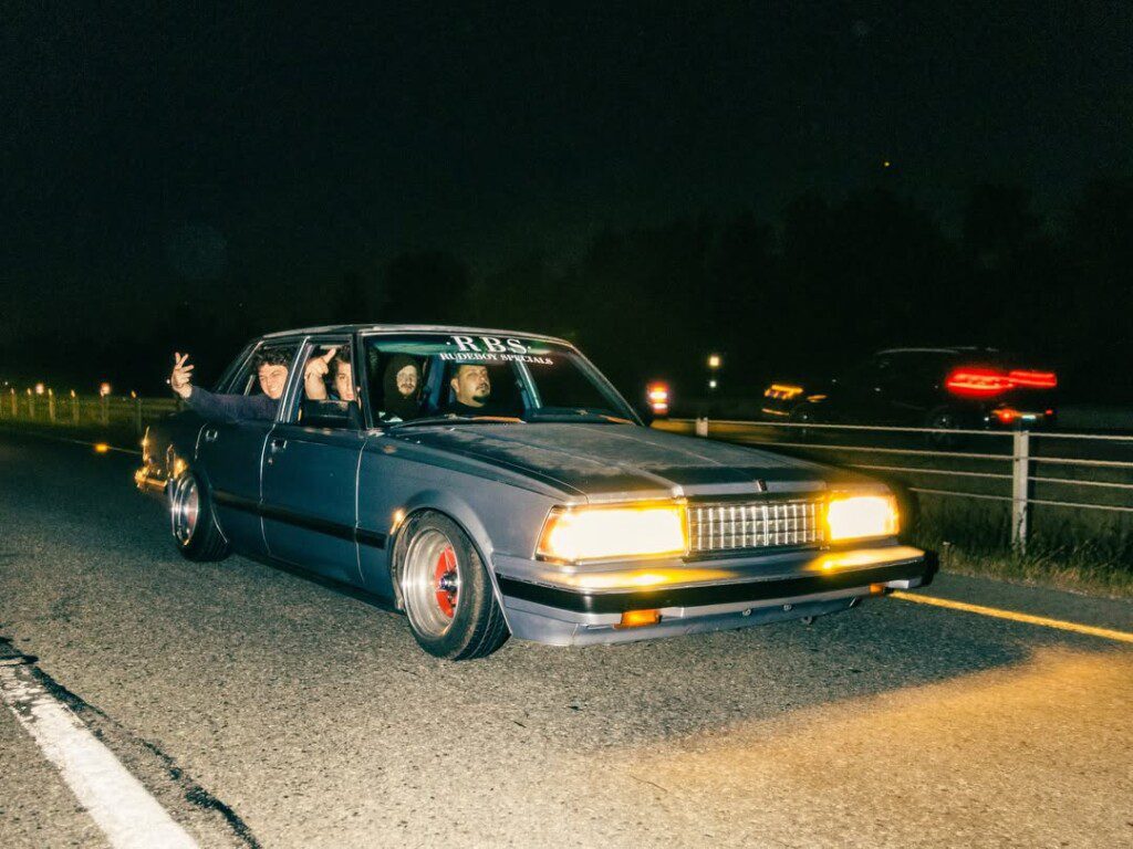 Vintage car cruising at night with passengers, glowing headlights, and blurred motion on a dimly lit highway.