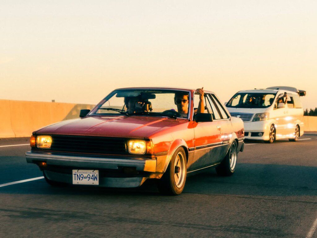 Red classic car driving on highway at sunset, leading a white van.