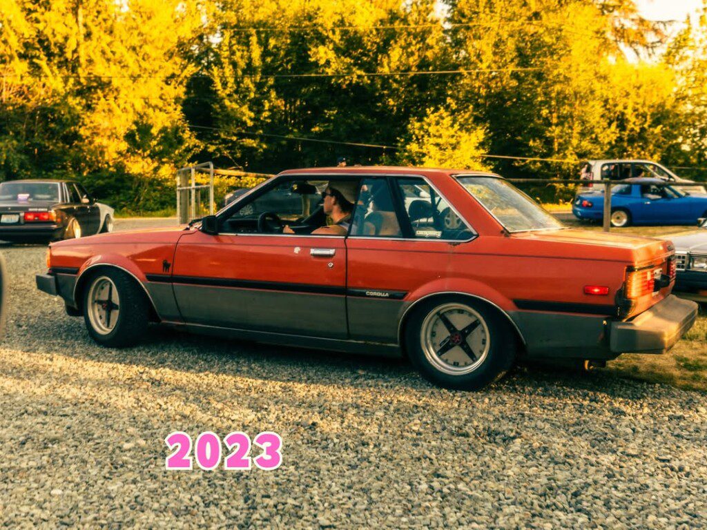 Vintage red Toyota Corolla parked on a gravel path at sunset with trees in the background.