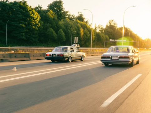Two vintage cars driving on a highway at sunset, surrounded by trees and bathed in warm sunlight.