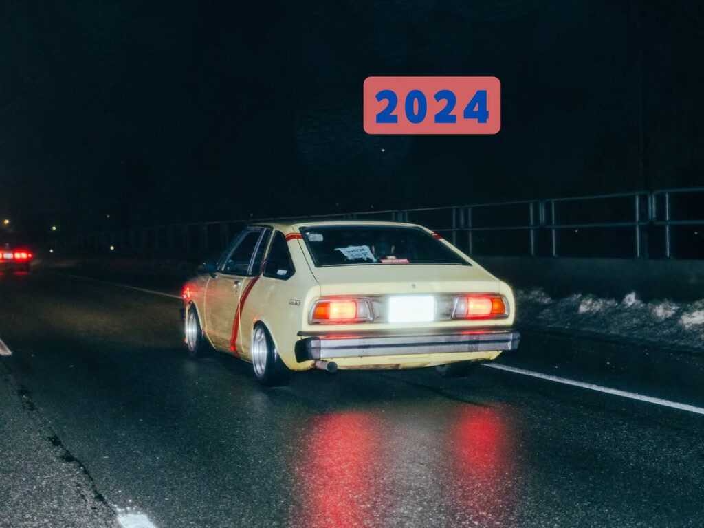 Vintage yellow car driving at night on wet road, featuring 2024 in bold text above.