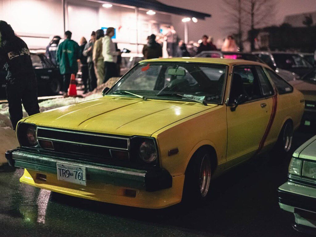 Vintage yellow car parked at a night car meet, surrounded by people and other vehicles.