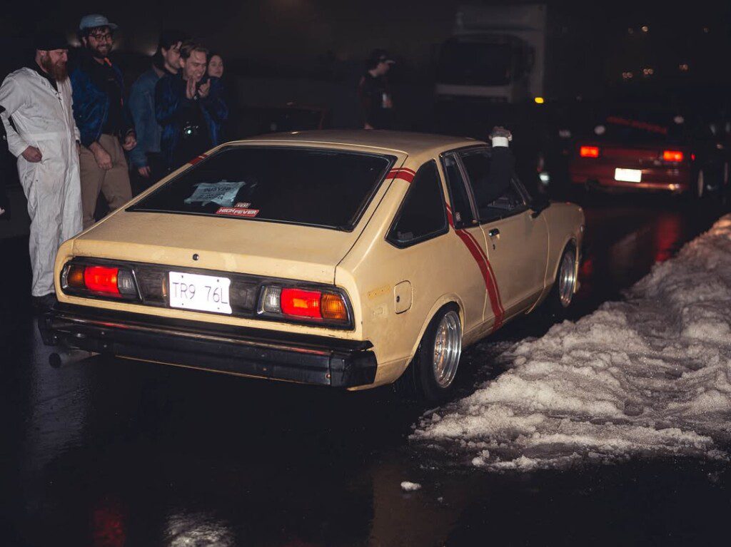 Vintage yellow car parked at night with spectators, snow nearby, creating a nostalgic automotive scene.