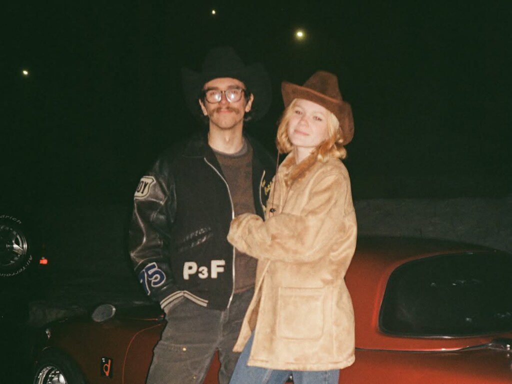 Two people in cowboy hats stand smiling at night in front of a vintage car.