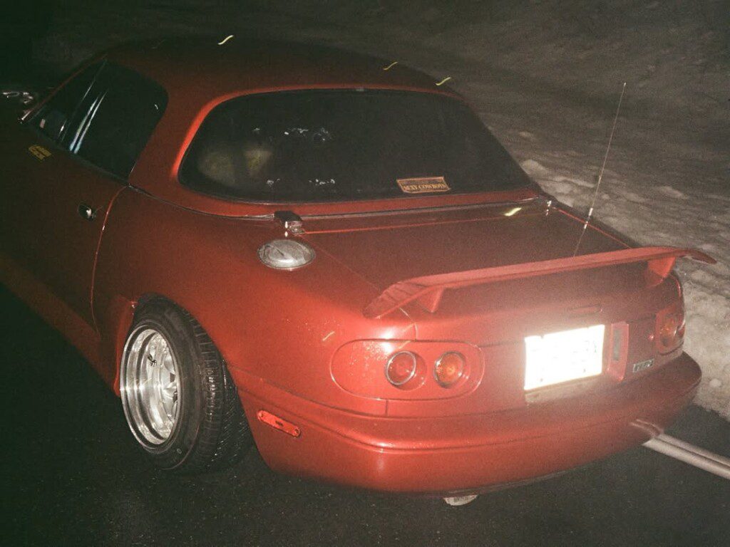 Red sports car with a spoiler parked at night on a wet road. Tail lights and custom wheels visible.
