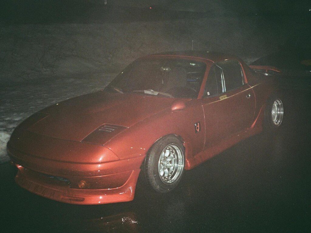 Red sports car parked on a wet road at night with snow in the background, showcasing sleek body and shiny wheels.