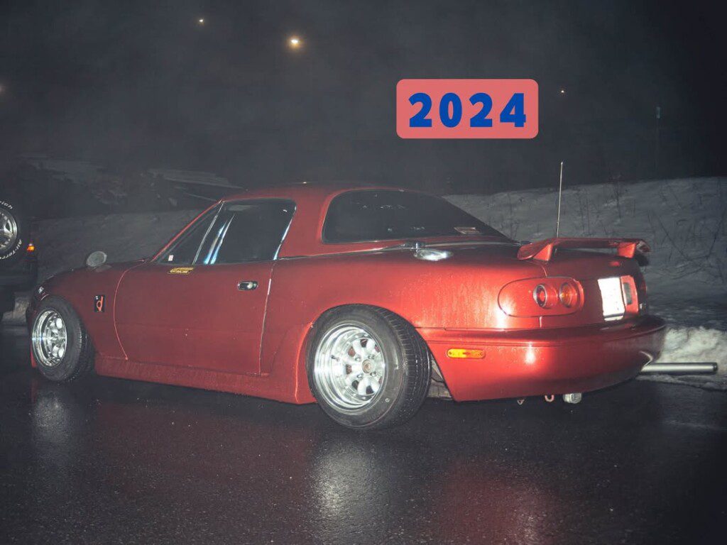 Red sports car parked at night on wet pavement with 2024 in blue on red above it, suggesting a future event.