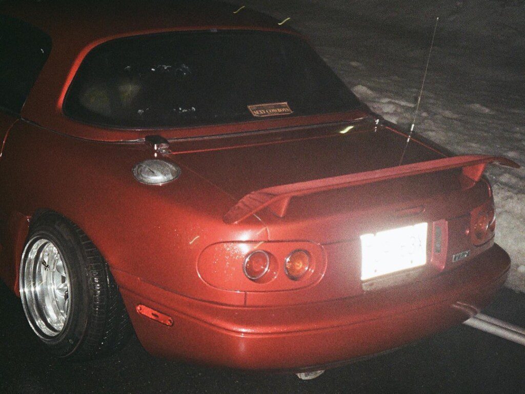Red sports car with rear spoiler and custom wheels parked at night on wet road, showcasing sleek design and style.