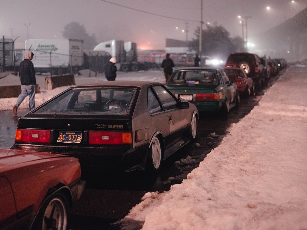 Row of vintage cars parked on a snowy street at night, with misty lighting and people walking nearby.