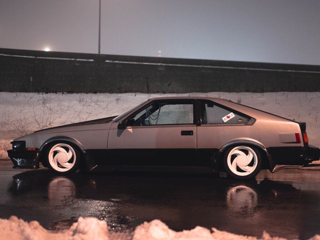 Side view of a vintage sports car with custom rims parked on a wet, snowy street at night.
