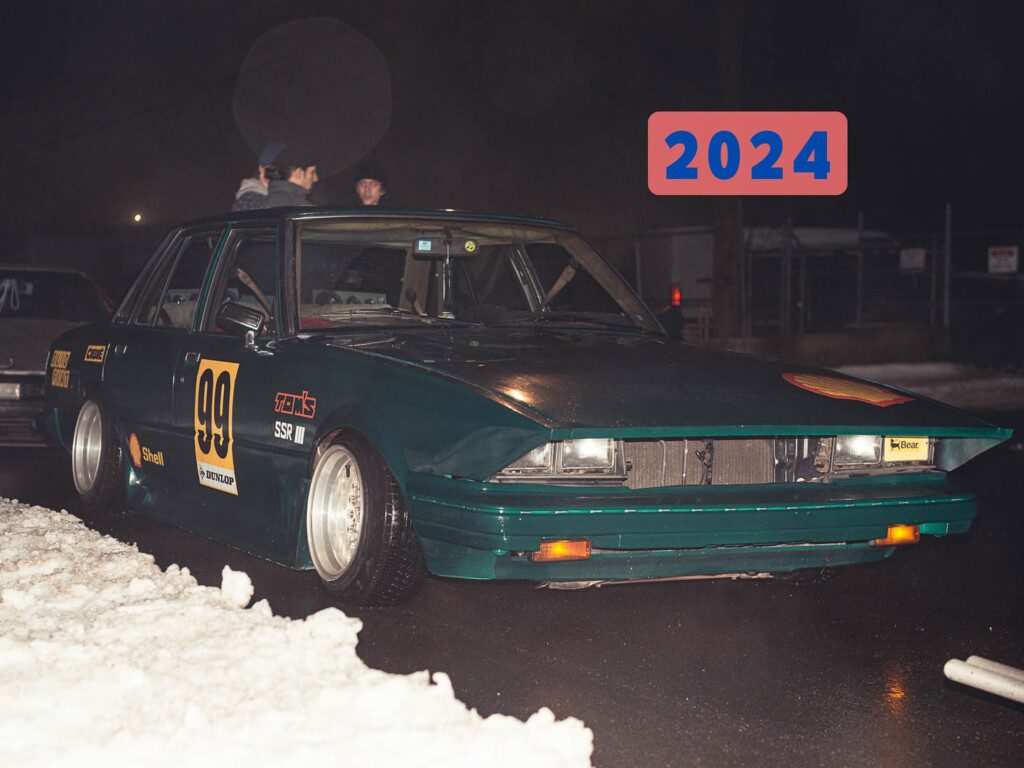 Vintage race car number 99 on wet track at night, 2024 event. Classic design with sponsors visible.