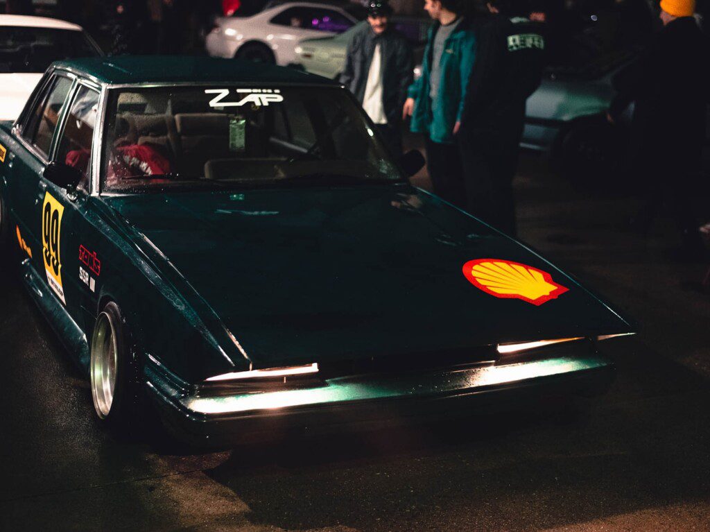Vintage racing car at night event with Shell logo, number 99, and a crowd in background. Retro car enthusiasts gathering.