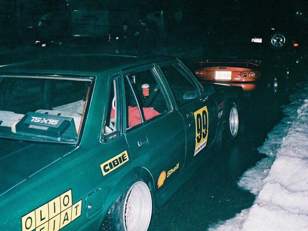 Vintage race cars parked on a snowy street at night, showcasing number 99 with racing decals.