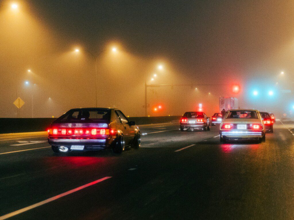 Vintage cars driving at night on a foggy road, illuminated by streetlights and taillights.