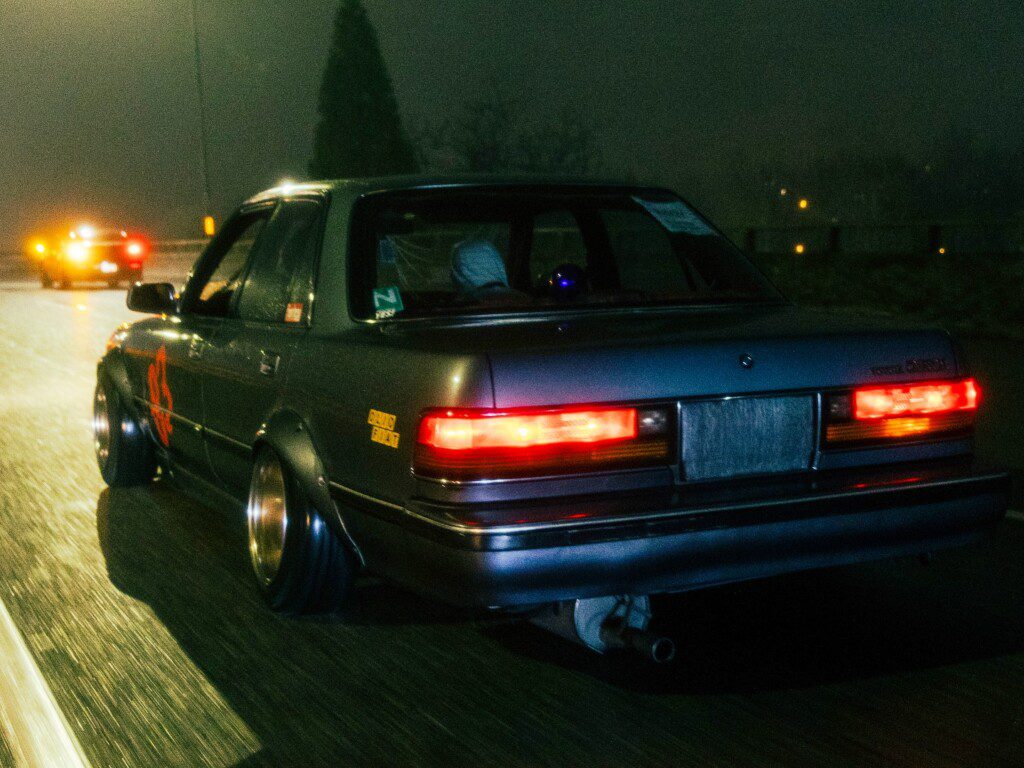 Vintage car with bright taillights driving on a misty night highway, styled with decals and wide wheels.
