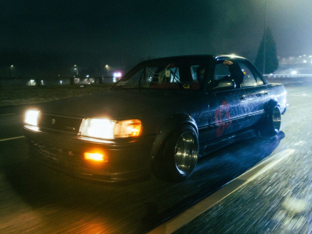 Vintage car driving in the rain at night with headlights on, creating a dynamic urban scene.