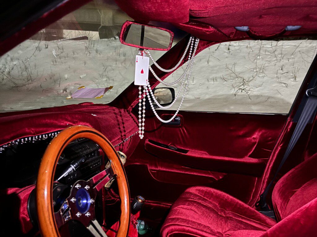 Vintage car interior with red velvet upholstery, wooden steering wheel, and hanging accessories including a playing card.