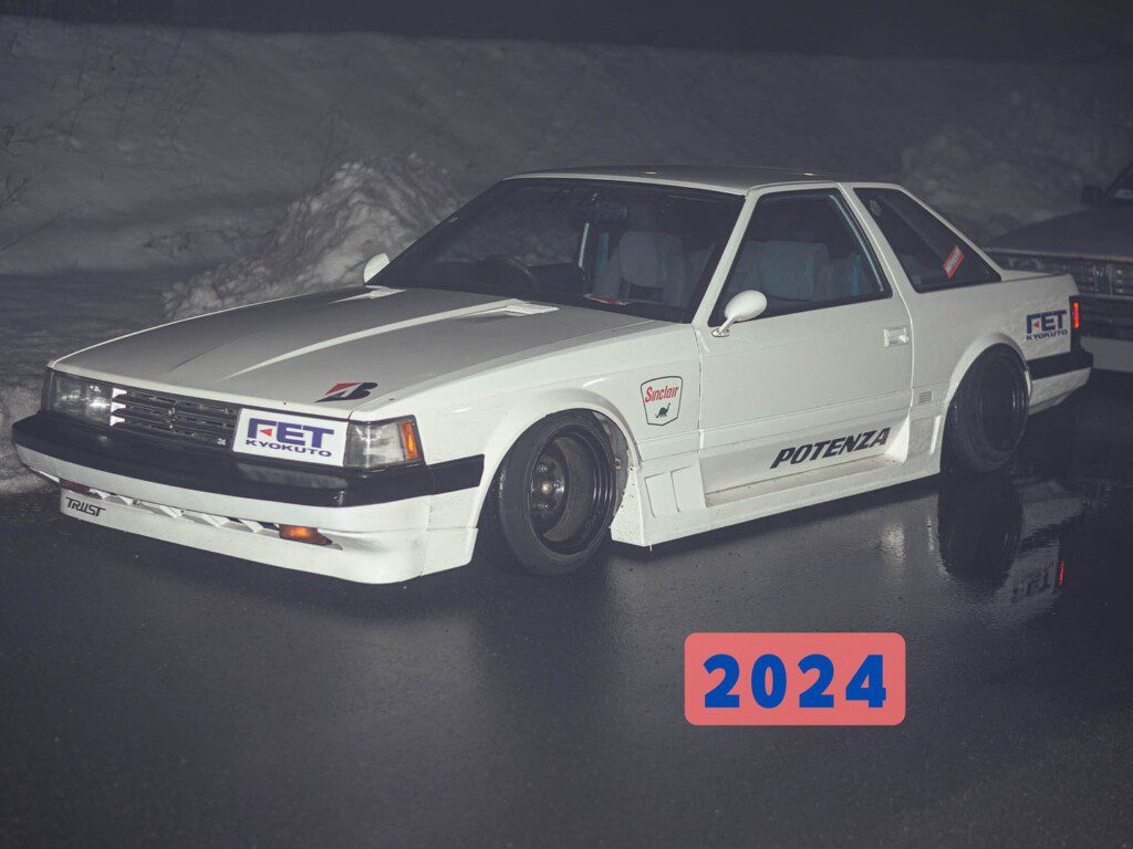 Modified white vintage car with decals, parked at night on reflective wet pavement; 2024 text overlay.